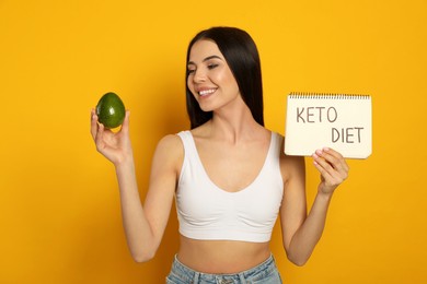 Photo of Happy woman holding avocado and notebook with words Keto Diet on yellow background