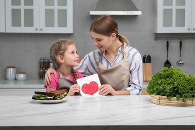 Little daughter congratulating mom with greeting card in kitchen. Happy Mother's Day