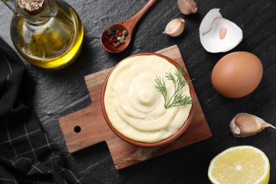 Photo of Fresh mayonnaise sauce in bowl and ingredients on black table, flat lay