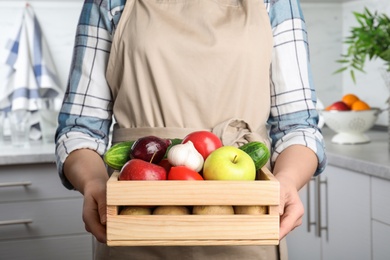 Photo of Woman holding wooden crate filled with fresh vegetables and fruits in kitchen, closeup