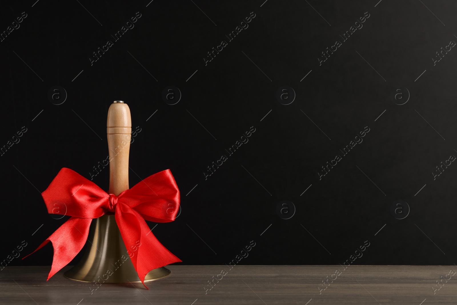 Photo of Golden bell with red bow on wooden table near blackboard, space for text. School days