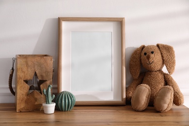 Photo of Empty photo frame near cute toy bunny and decor on wooden table, space for text. Baby room interior element