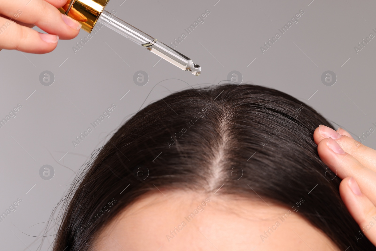 Photo of Woman applying essential oil onto hair roots on light grey background, closeup