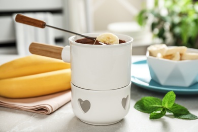 Photo of Dipping slice of banana into fondue pot with milk chocolate on table indoors