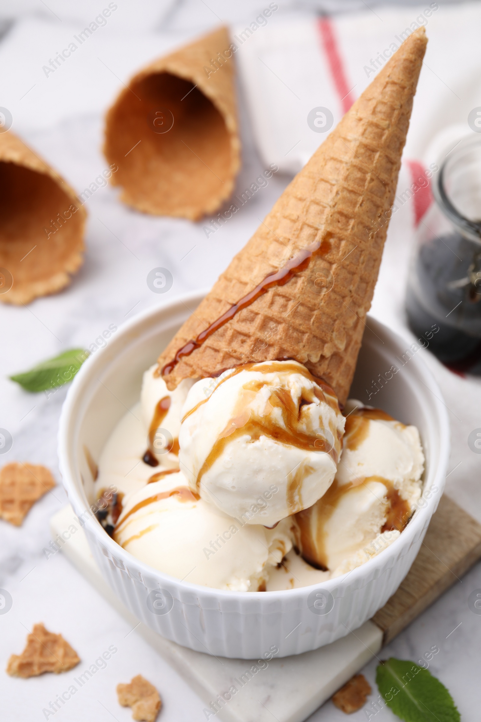 Photo of Scoops of ice cream with caramel sauce and wafer cone on white marble table, closeup