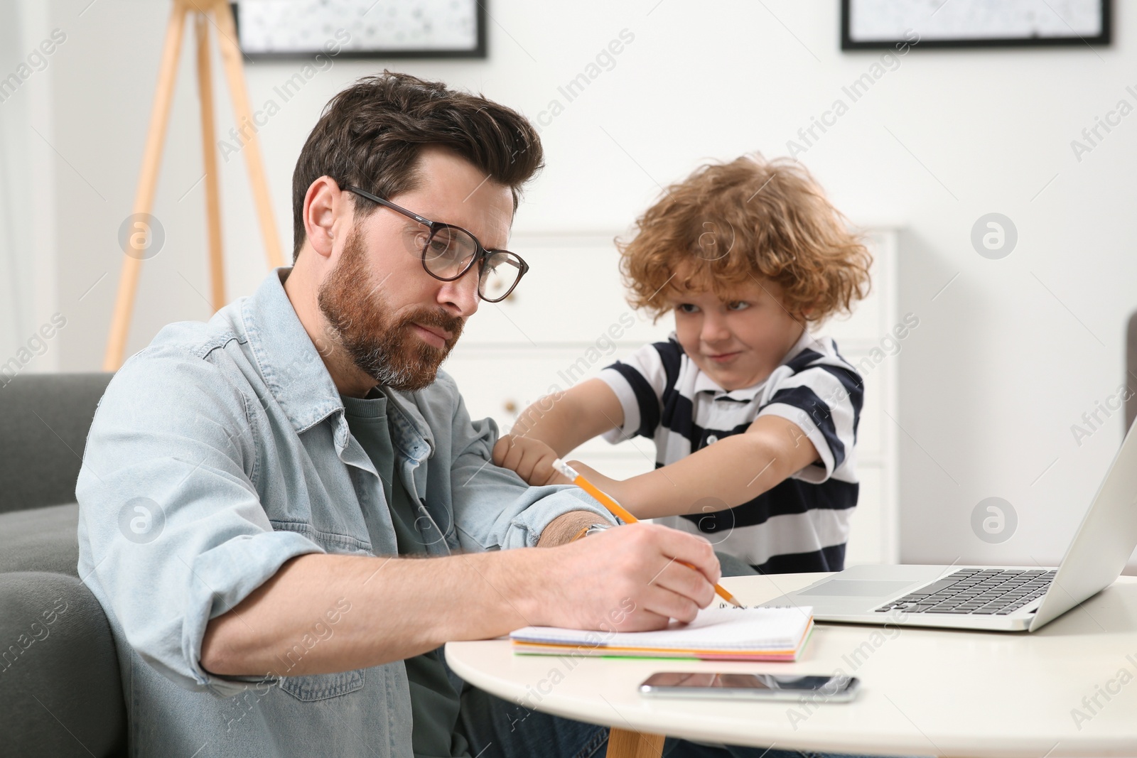 Photo of Little boy bothering his father at home. Man working remotely at desk