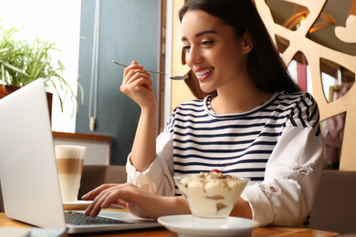 Young blogger with laptop eating dessert in cafe