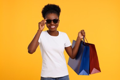 Photo of Happy young woman in stylish sunglasses with shopping bags on orange background