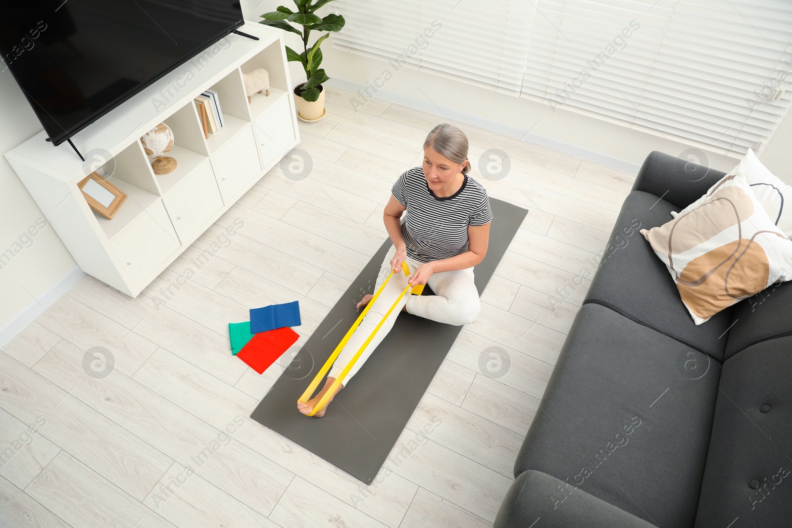 Photo of Senior woman doing exercise with fitness elastic band on mat at home, above view