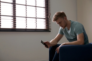 Lonely man with mobile phone in armchair near window indoors
