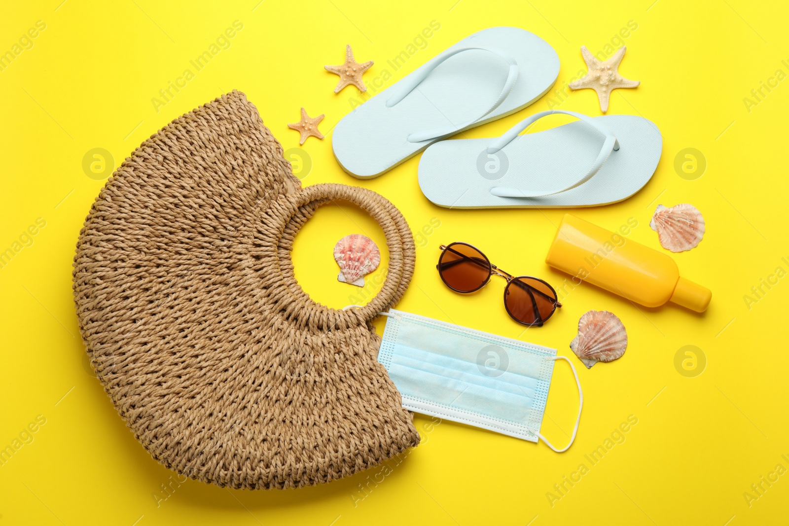 Photo of Stylish bag with beach accessories and protective mask on yellow background, flat lay