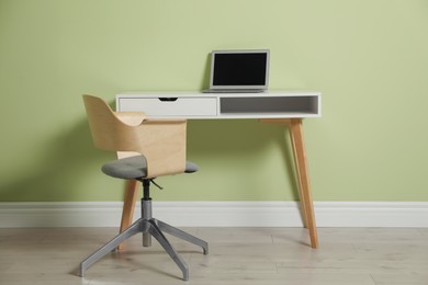 Stylish workplace with laptop and comfortable chair near green wall indoors. Interior design