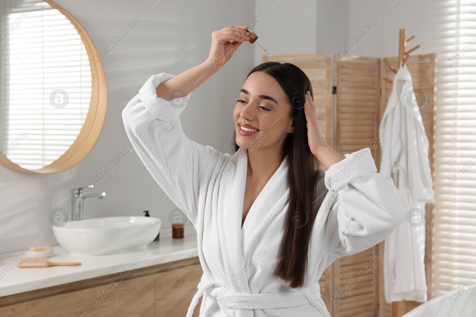 Photo of Happy young woman applying essential oil onto hair roots in bathroom