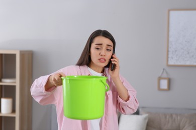 Photo of Young woman calling roof repair service while collecting leaking water from ceiling at home