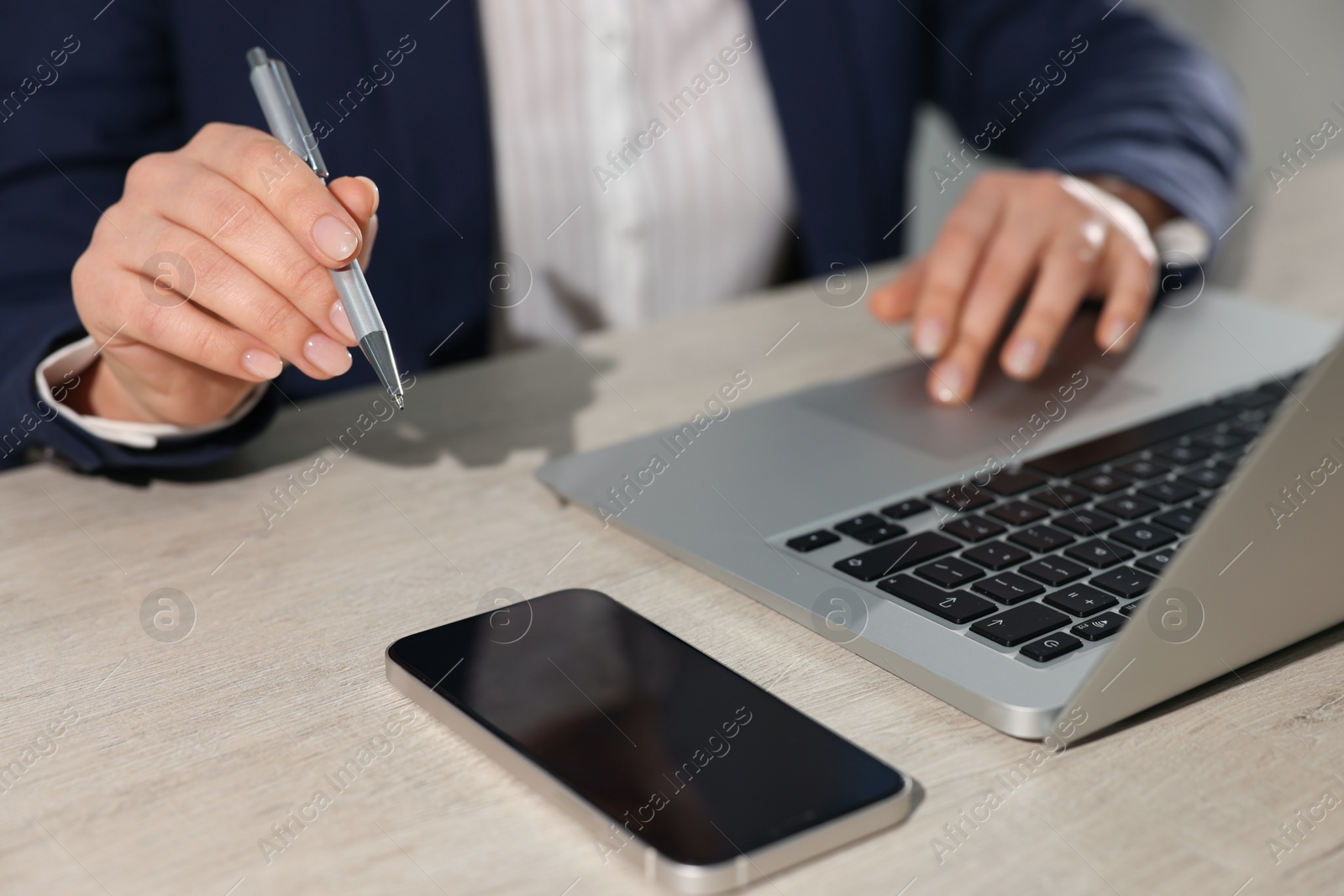 Photo of Woman with smartphone and pen working on laptop at wooden table, closeup. Electronic document management