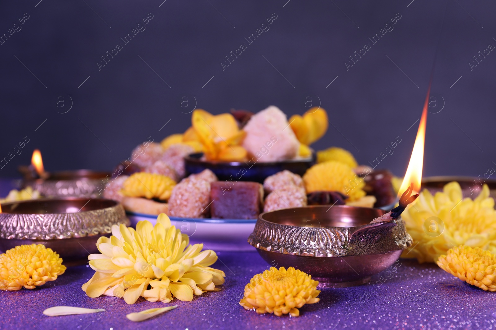 Photo of Diwali celebration. Diya lamps, tasty Indian sweets and chrysanthemum flowers on shiny violet table