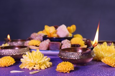 Diwali celebration. Diya lamps, tasty Indian sweets and chrysanthemum flowers on shiny violet table