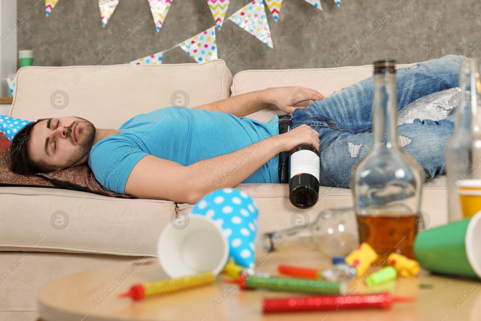 Photo of Young man with bottle of wine sleeping on sofa in messy room after party