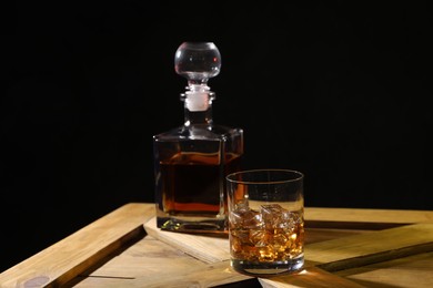 Whiskey with ice cubes in glass and bottle on wooden crate against black background