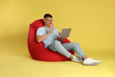 Photo of Handsome man with laptop waving hello on red bean bag chair against yellow background