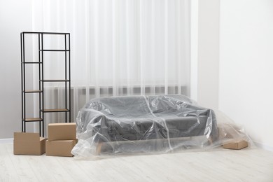 Photo of Stylish sofa covered with plastic film, shelving unit and boxes at home