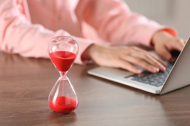 Hourglass with flowing sand on wooden table, selective focus. Man using laptop indoors