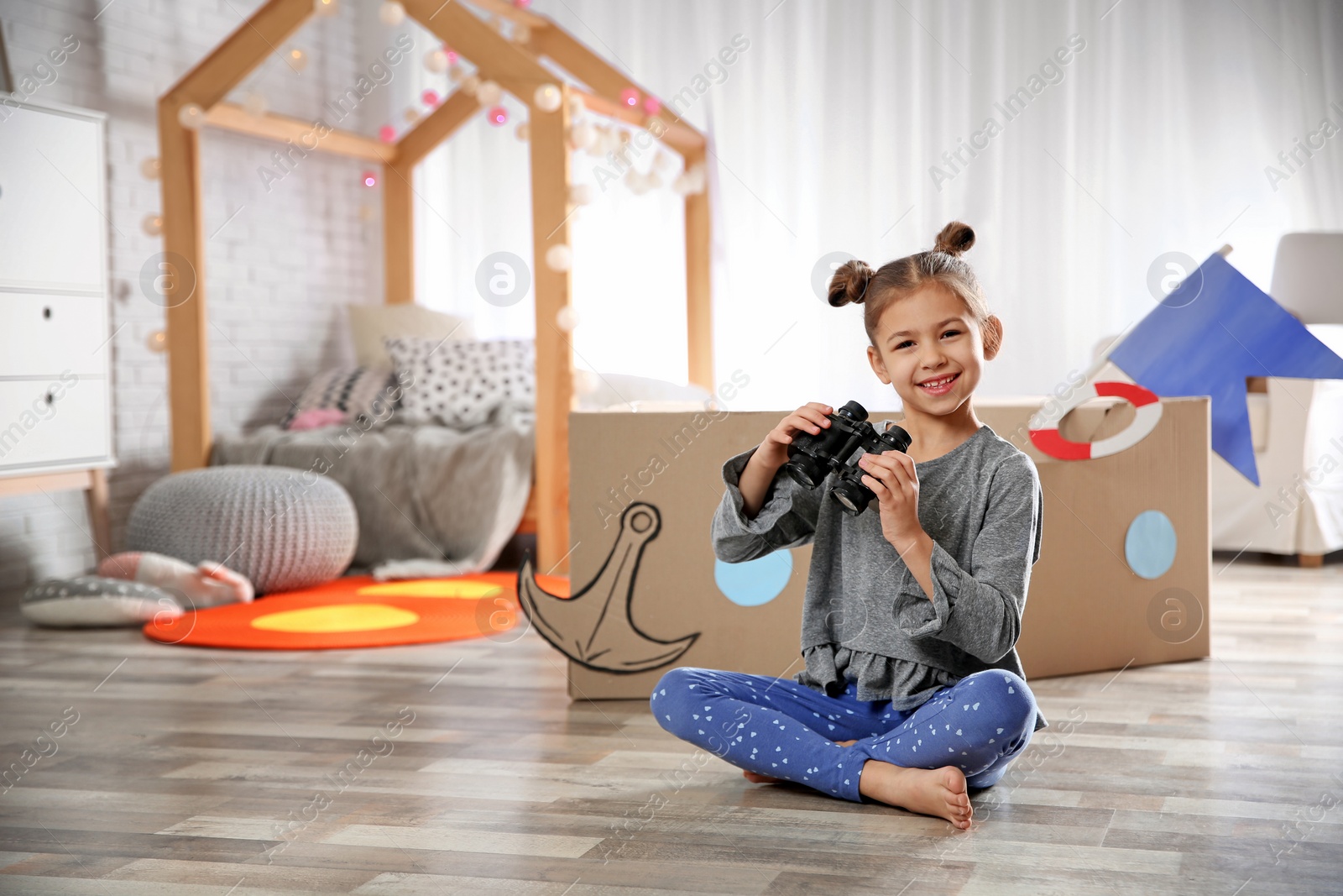 Photo of Cute little girl playing with binoculars and cardboard boat in bedroom