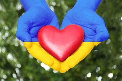 Image of Man with hands in colors of Ukrainian flag holding red heart outdoors, closeup. Volunteering during war