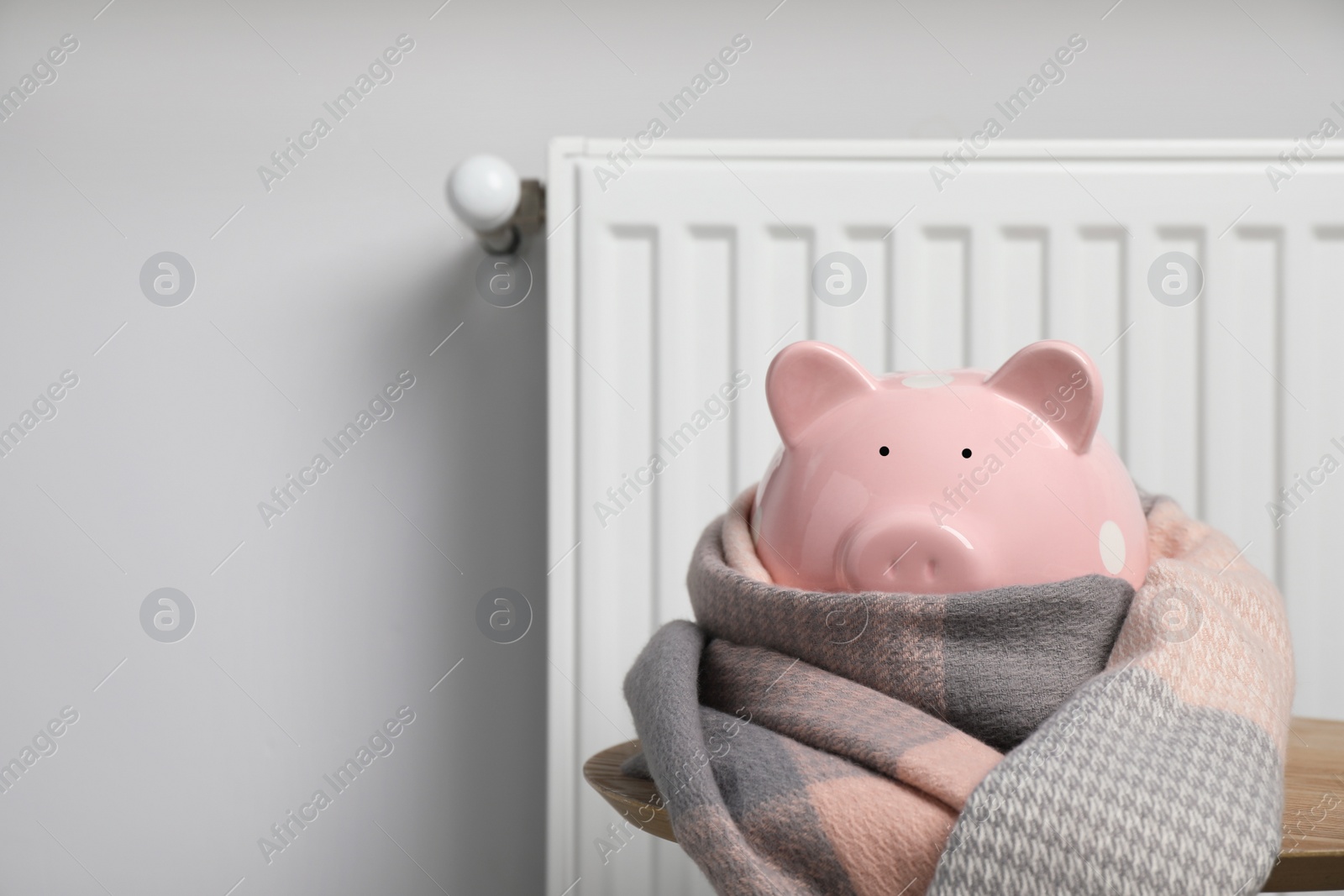 Photo of Piggy bank wrapped in scarf on table near heating radiator, space for text