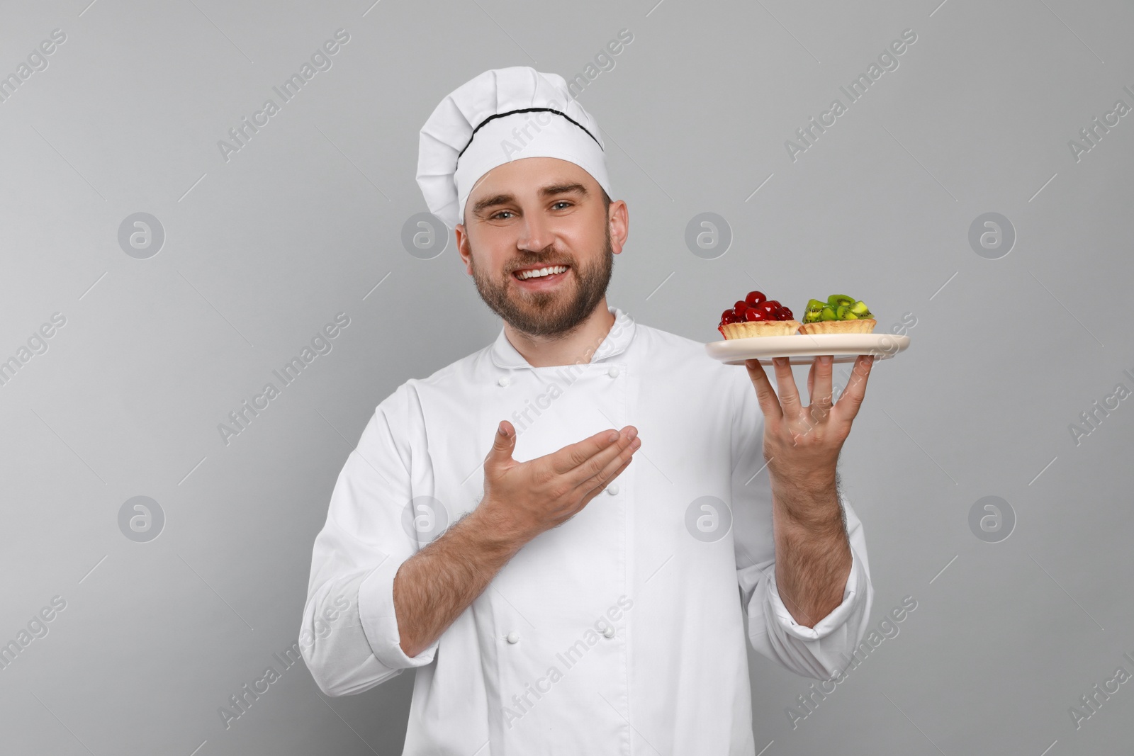 Photo of Happy professional confectioner in uniform holding plate with delicious tartlets on light grey background