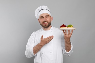 Happy professional confectioner in uniform holding plate with delicious tartlets on light grey background