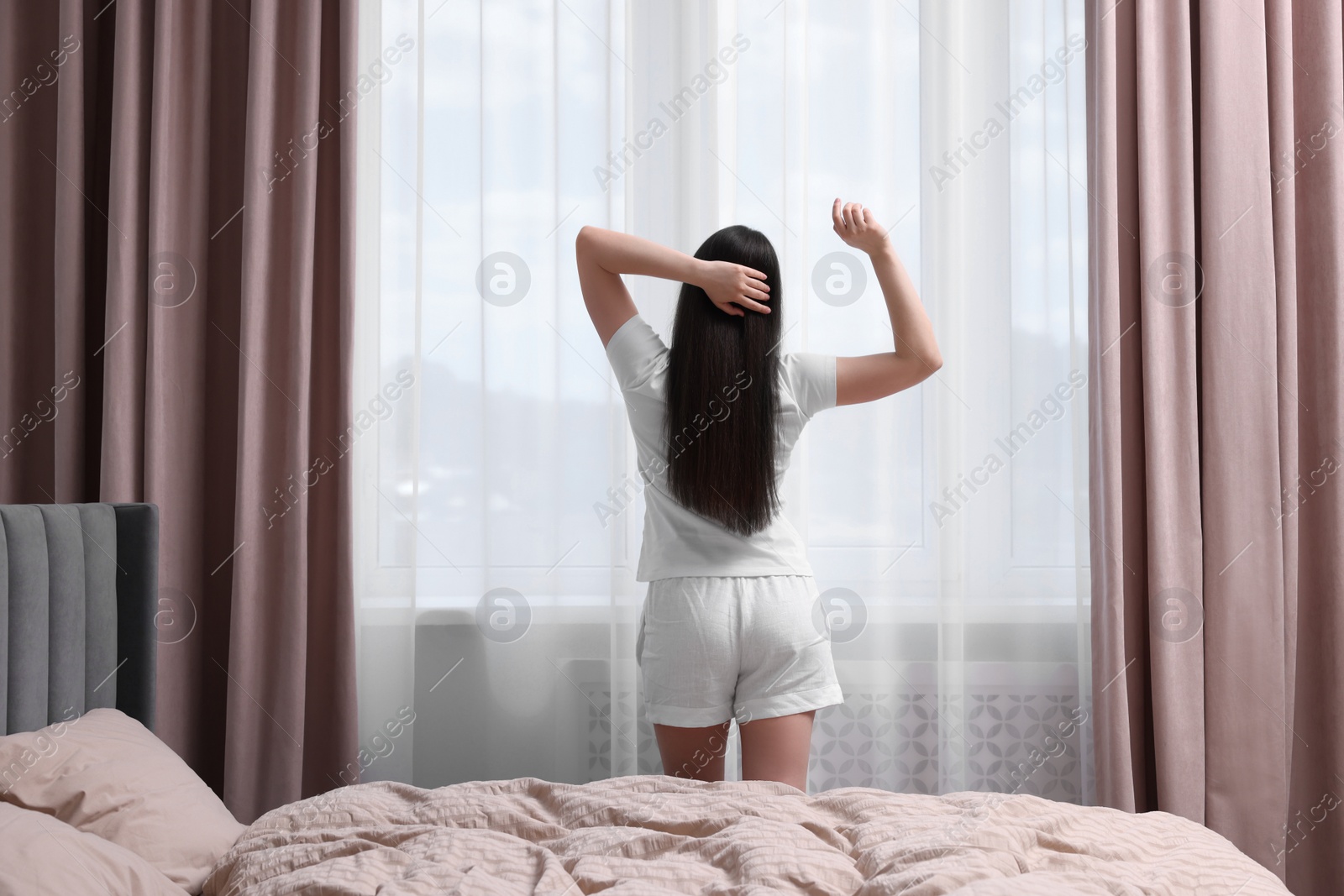 Photo of Woman stretching near window in bedroom, back view. Lazy morning