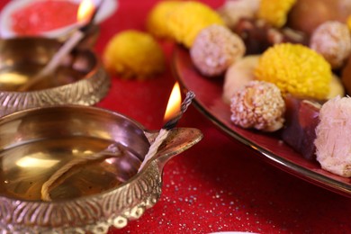 Diwali celebration. Diya lamps and tasty Indian sweets on shiny red table, closeup