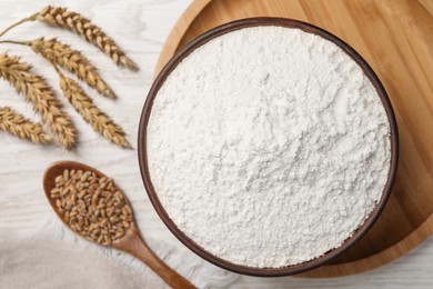 Organic flour in bowl, spoon with grains and ears of wheat on white wooden table, flat lay