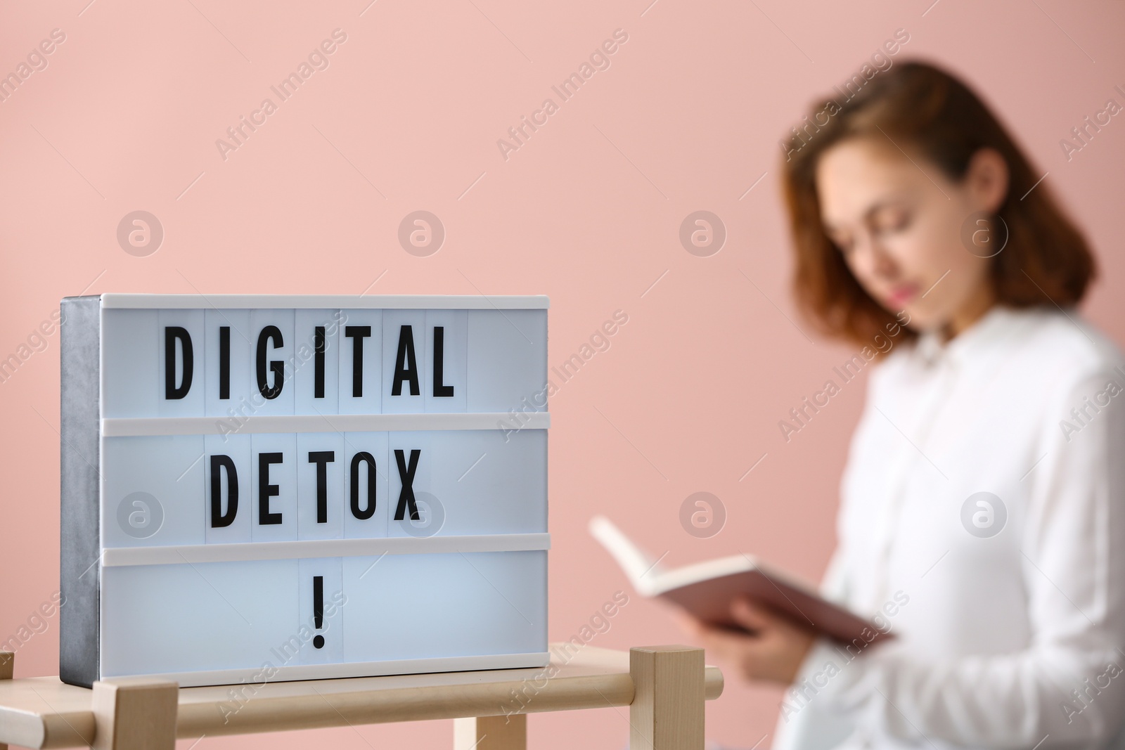 Photo of Woman reading book against pink background, focus on lightbox with phrase DIGITAL DETOX