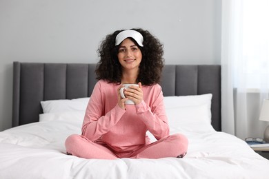 Photo of Beautiful young woman in stylish pyjama and sleep mask with cup of drink on bed at home