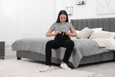 Woman trying to put on tight jeans on bed at home