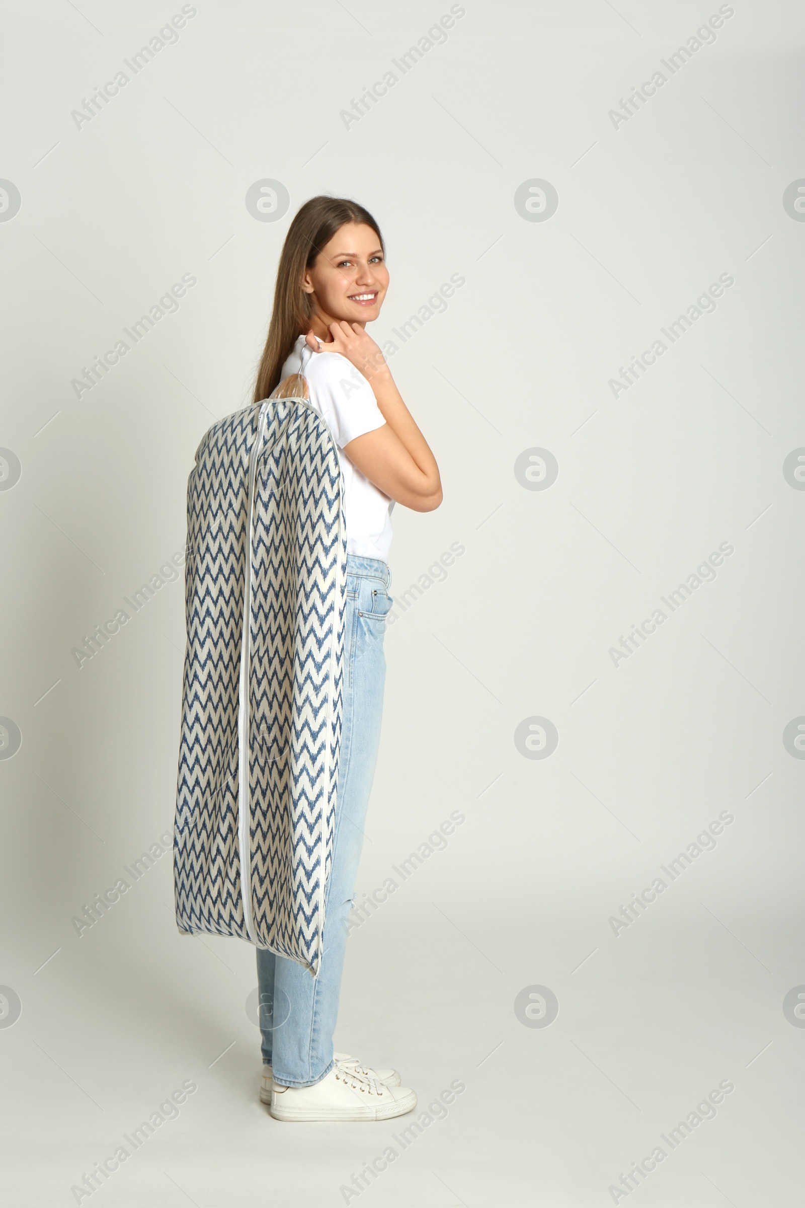 Photo of Young woman holding hanger with clothes in garment cover on light grey background, space for text. Dry-cleaning service