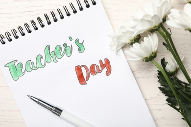 Photo of Notebook with words TEACHER'S DAY, pen and flowers on white wooden table, flat lay