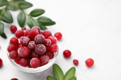 Frozen red cranberries in bowl and green leaves on white table, closeup. Space for text