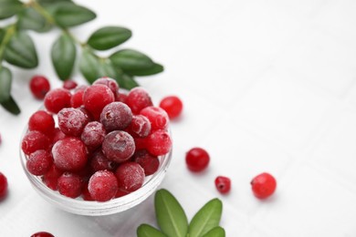 Photo of Frozen red cranberries in bowl and green leaves on white table, closeup. Space for text