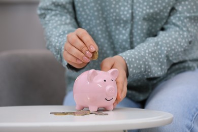 Photo of Young woman putting coin into piggy bank at table indoors, closeup