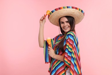 Photo of Young woman in Mexican sombrero hat and poncho dancing with maracas on pink background