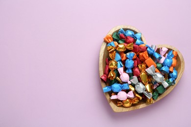 Photo of Heart shaped plate with candies in colorful wrappers on violet background, top view. Space for text