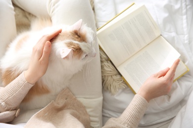 Woman with cute fluffy cat and book on bed, top view