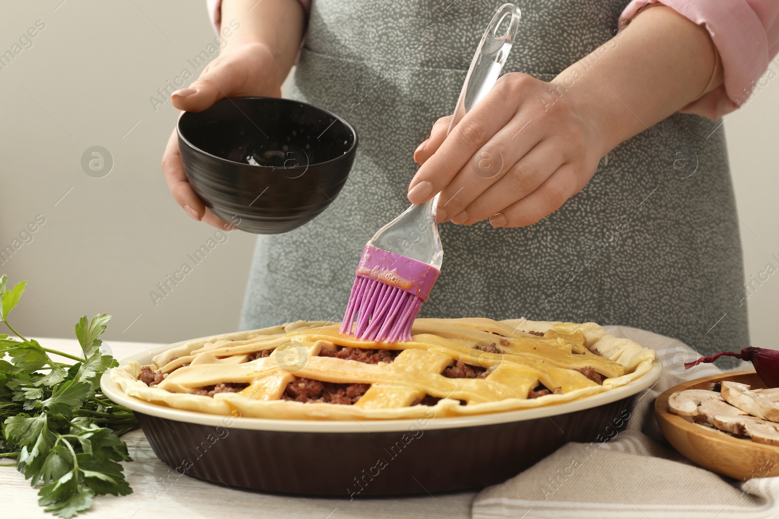 Photo of Woman spreading egg yolk onto raw meat pie at white wooden table, closeup