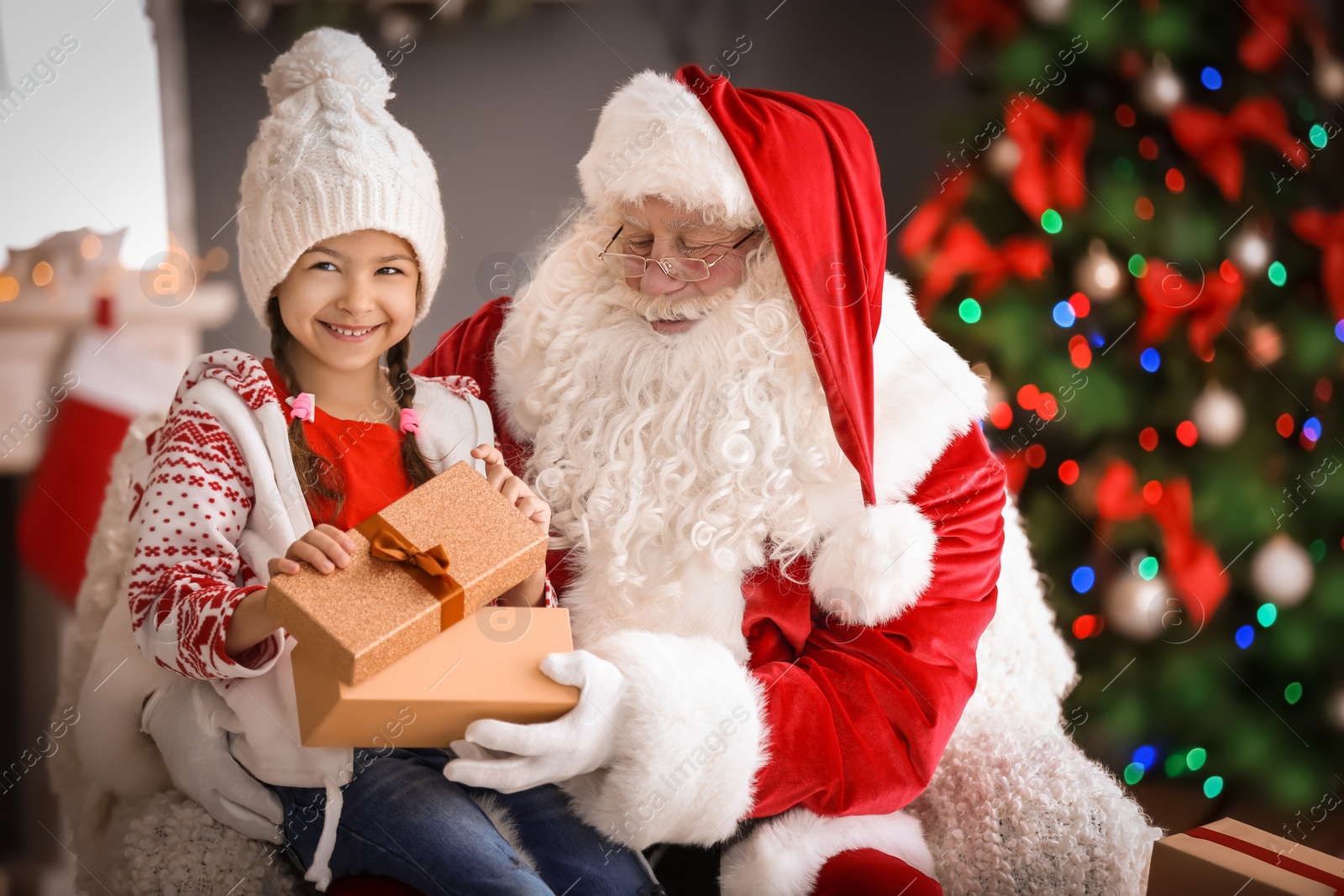 Photo of Little girl with gift box sitting on authentic Santa Claus' lap indoors