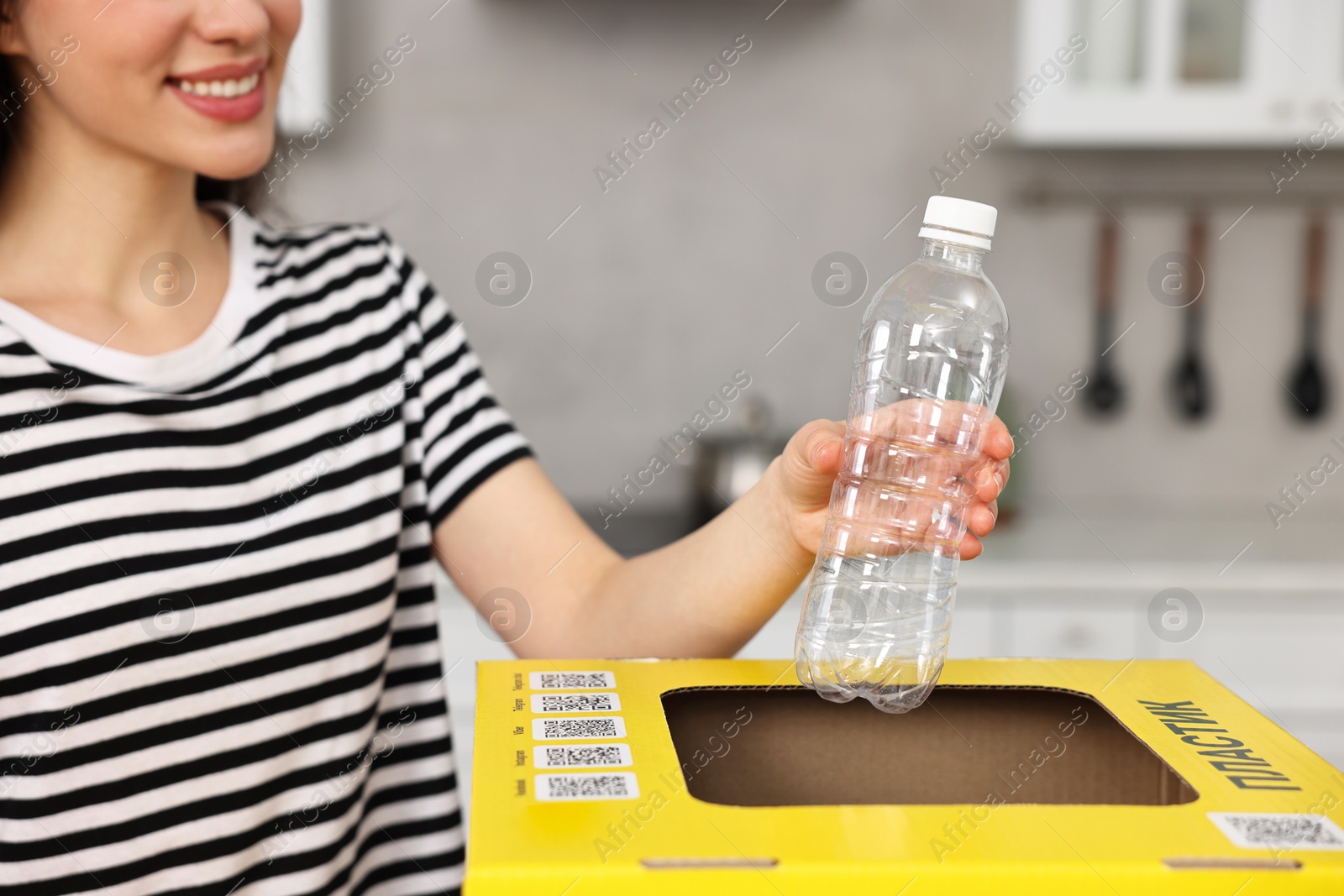 Photo of Garbage sorting. Smiling woman throwing plastic container into cardboard box in kitchen, closeup