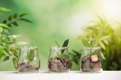 Glass jars of coins with young plant on white table against blurred background. Space for text