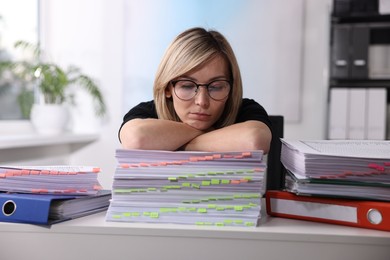 Overwhelmed woman sitting at table with stacks of documents and folders in office